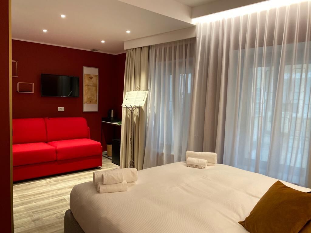 Habitación de hotel con una cama grande, un sofá rojo, un televisor de pantalla plana y una ventana con cortinas translúcidas.