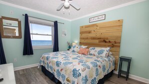 A bedroom with a wooden headboard, a bed with floral bedding, a window with curtains, and a small table. - OCEANFRONT, Community Pool and beach walkway! KD220, Feelin Nauti (Kill Devil Hills)
