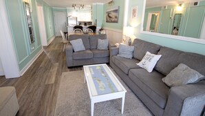 A living room with a grey sofa, a white coffee table, and a kitchen in the background. - OCEANFRONT, Community Pool and beach walkway! KD220, Feelin Nauti (Kill Devil Hills)
