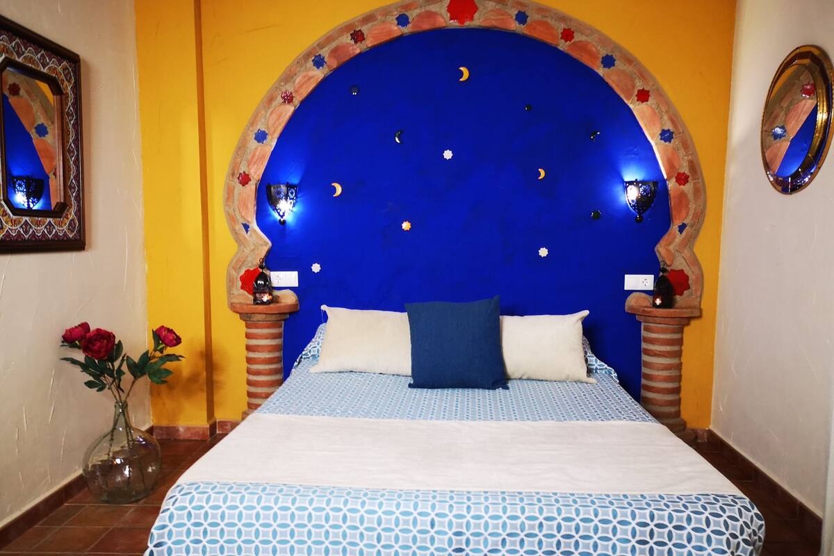 Ein Schlafzimmer mit blauem Kopfteil, einem Bett mit gemusterten Bettbezügen, einer Vase mit Blumen und einem Spiegel an der Wand.