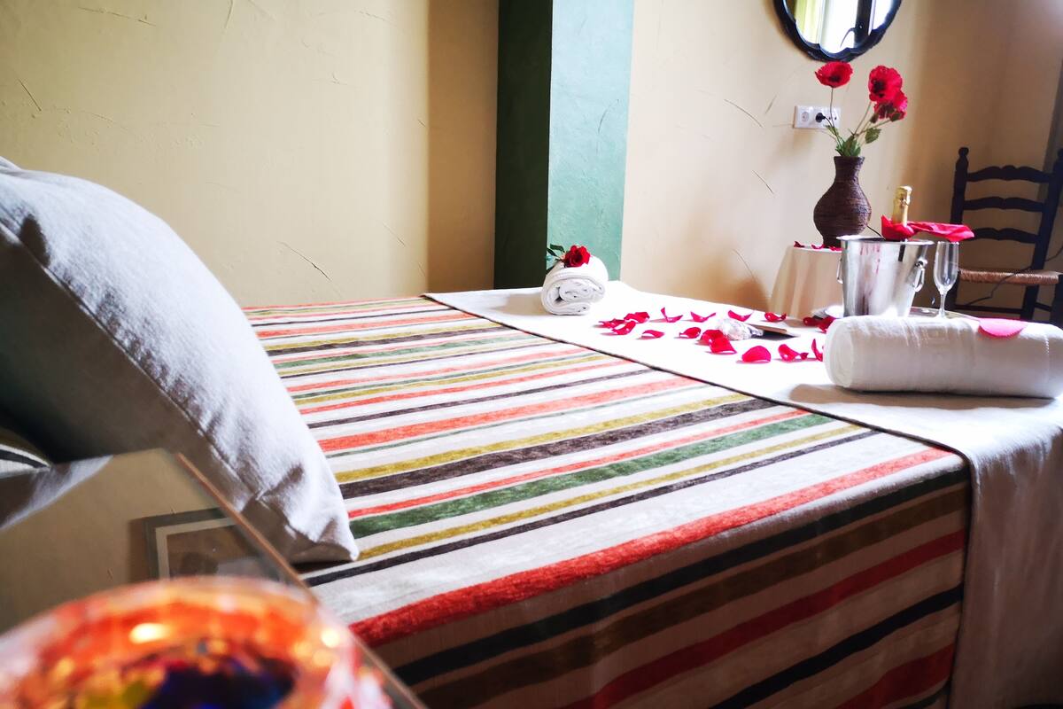 Ein Hotelzimmer mit gestreifter Bettdecke, einem ordentlich bezogenen Bett mit einem weißen Kopfkissen, einer Vase mit roten Blumen, einem kleinen Tisch mit einem Glas und einer Flasche sowie einem Stuhl.