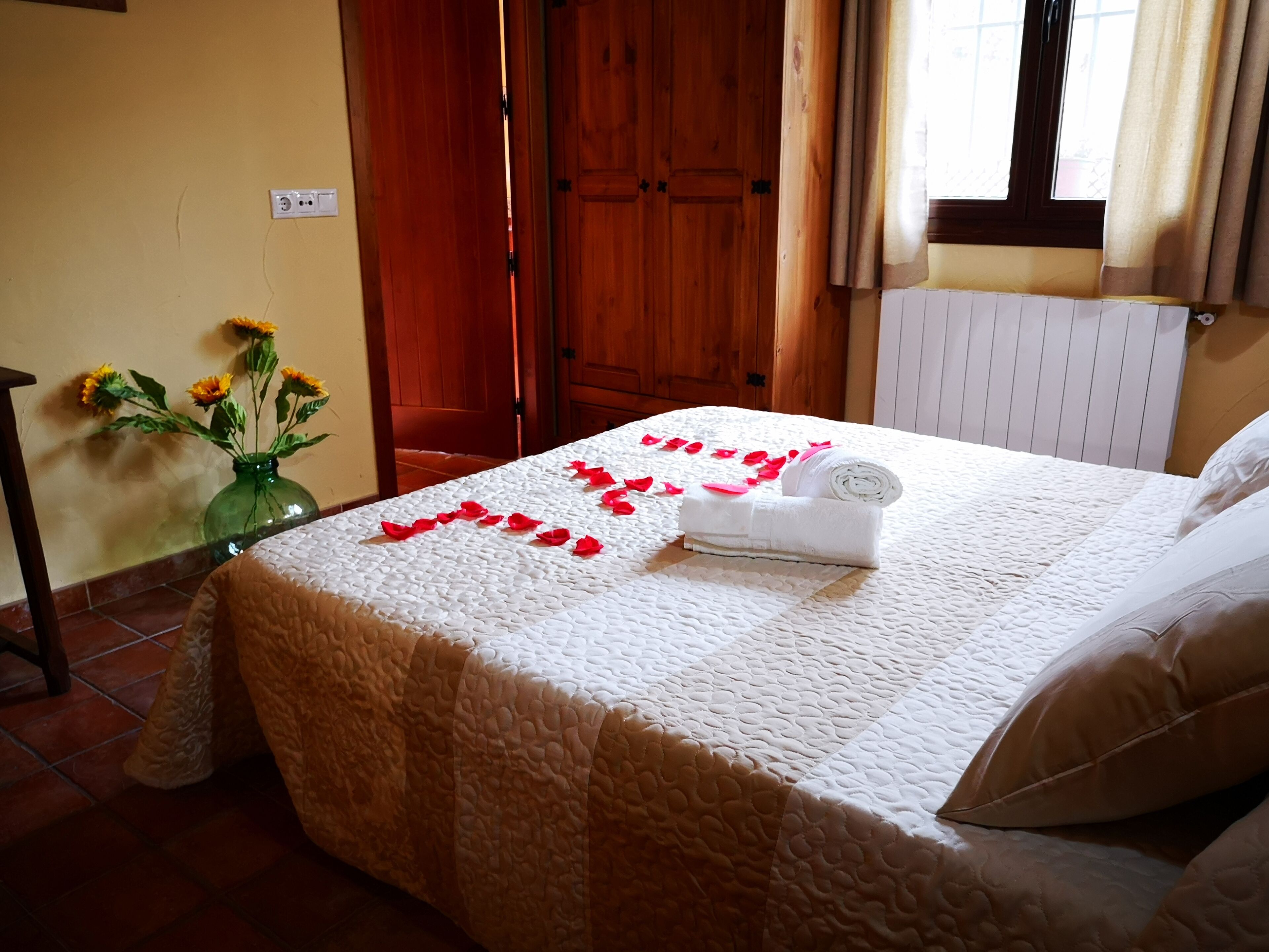 Una cama bien hecha con colcha blanca, una toalla enrollada y pétalos de rosa dispuestos sobre la cama.
