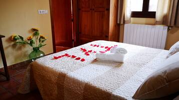 Una cama bien hecha con colcha blanca, una toalla enrollada y pétalos de rosa dispuestos sobre la cama.