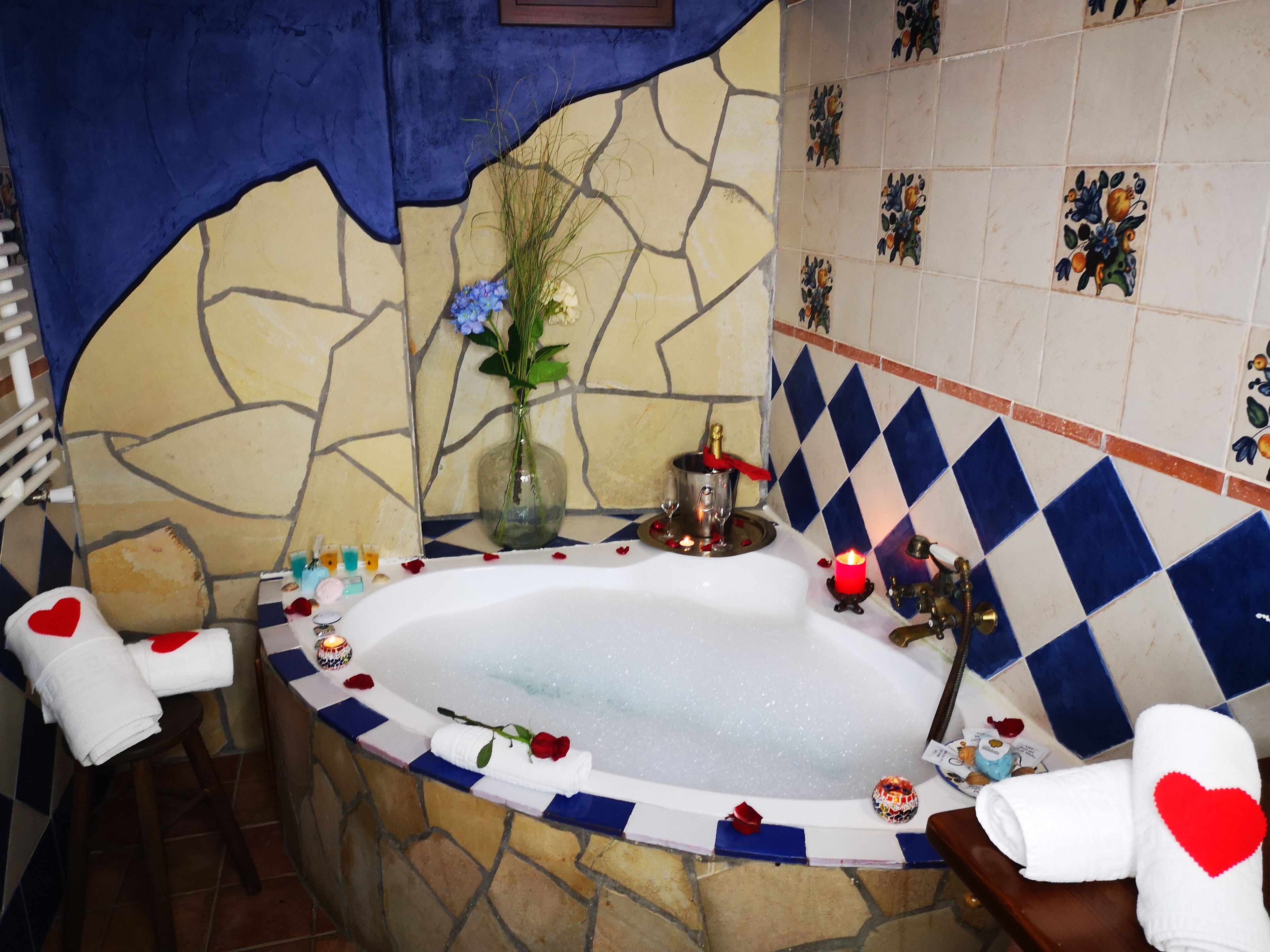 Une salle de bain avec un mur en pierre, une bordure de carreaux bleus et blancs, une baignoire remplie de pétales de roses, un vase avec des fleurs et des serviettes ornées de motifs en forme de cœur.