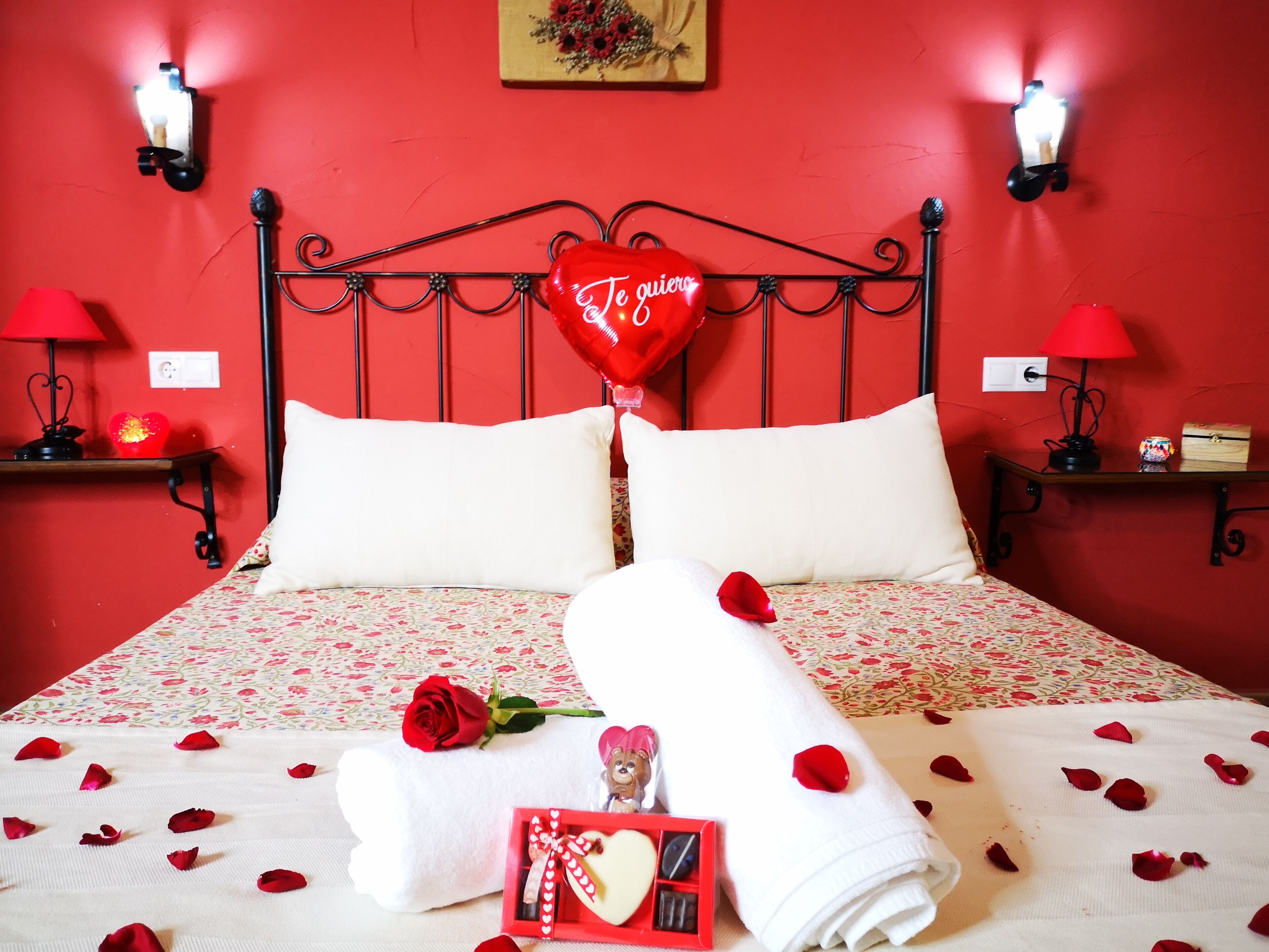 A red-themed bedroom with a bed, pillows, a heart-shaped balloon, rose petals, a rolled towel, a small figurine, and a red box with a heart design.
