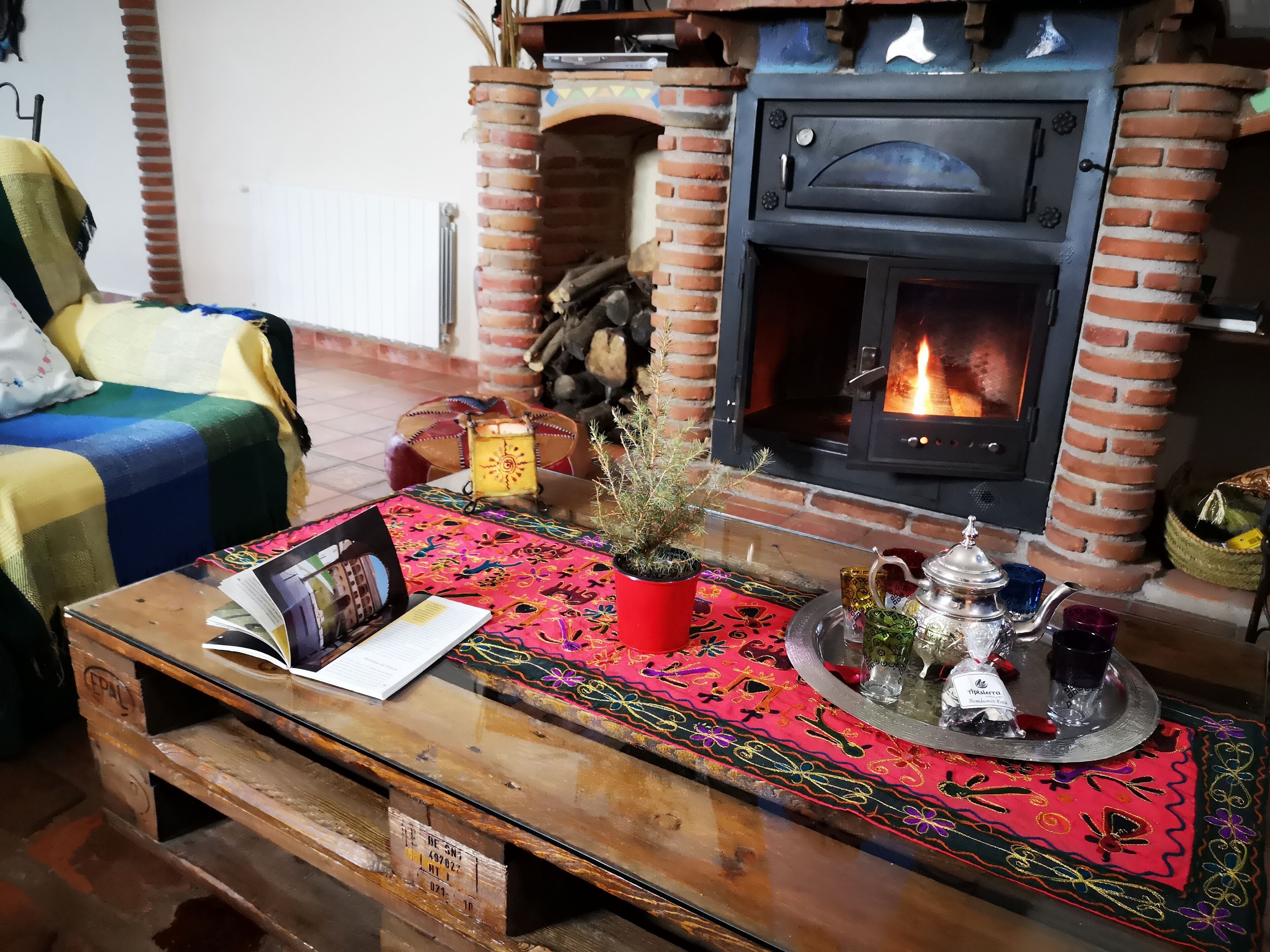 A cozy living room with a brick fireplace, a wooden coffee table with a colorful table runner, a potted plant, and a tea set.
