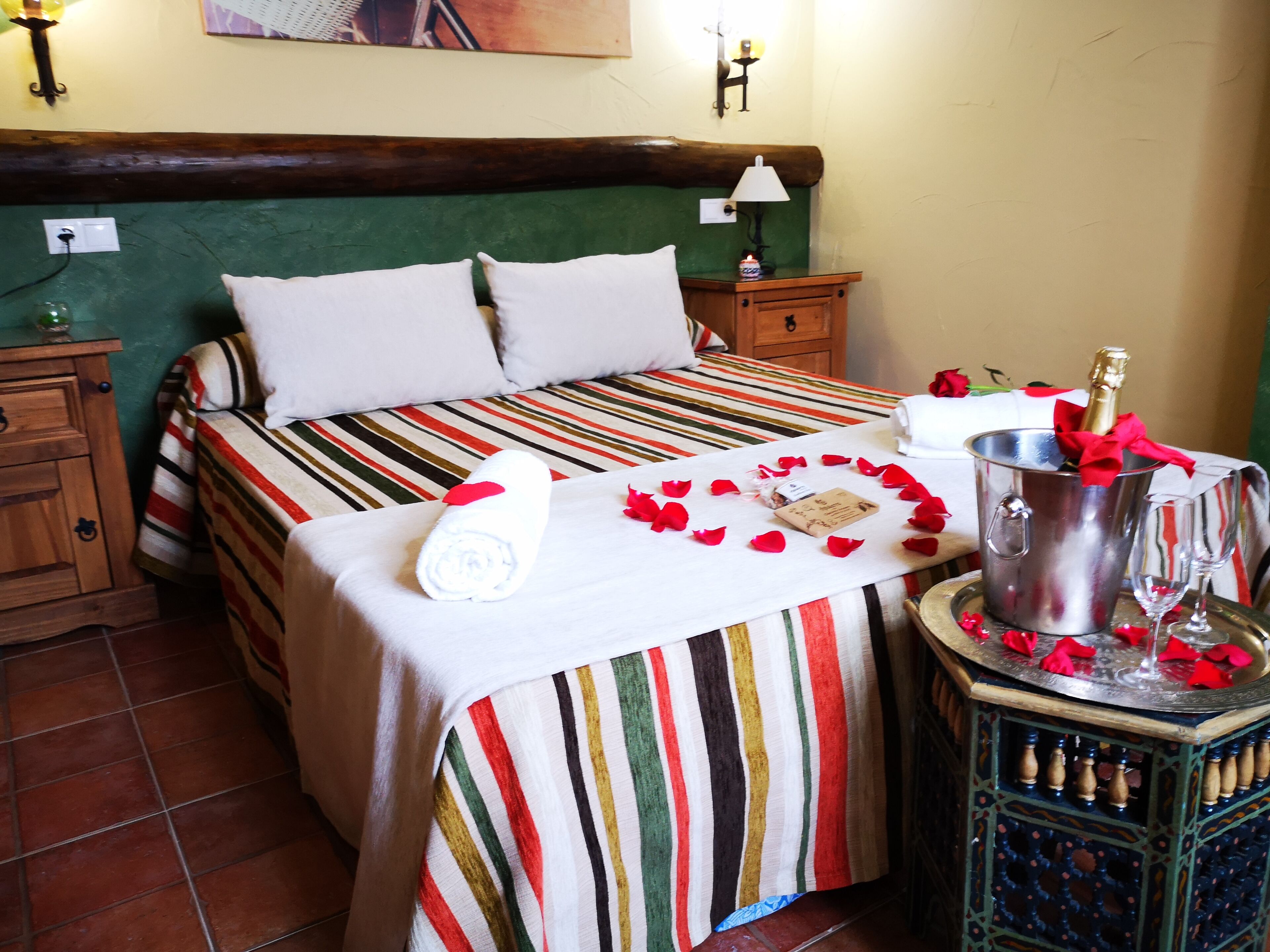 A neatly made bed with a striped bedspread, a bedside table with a bottle of champagne, and rose petals arranged on the bed.