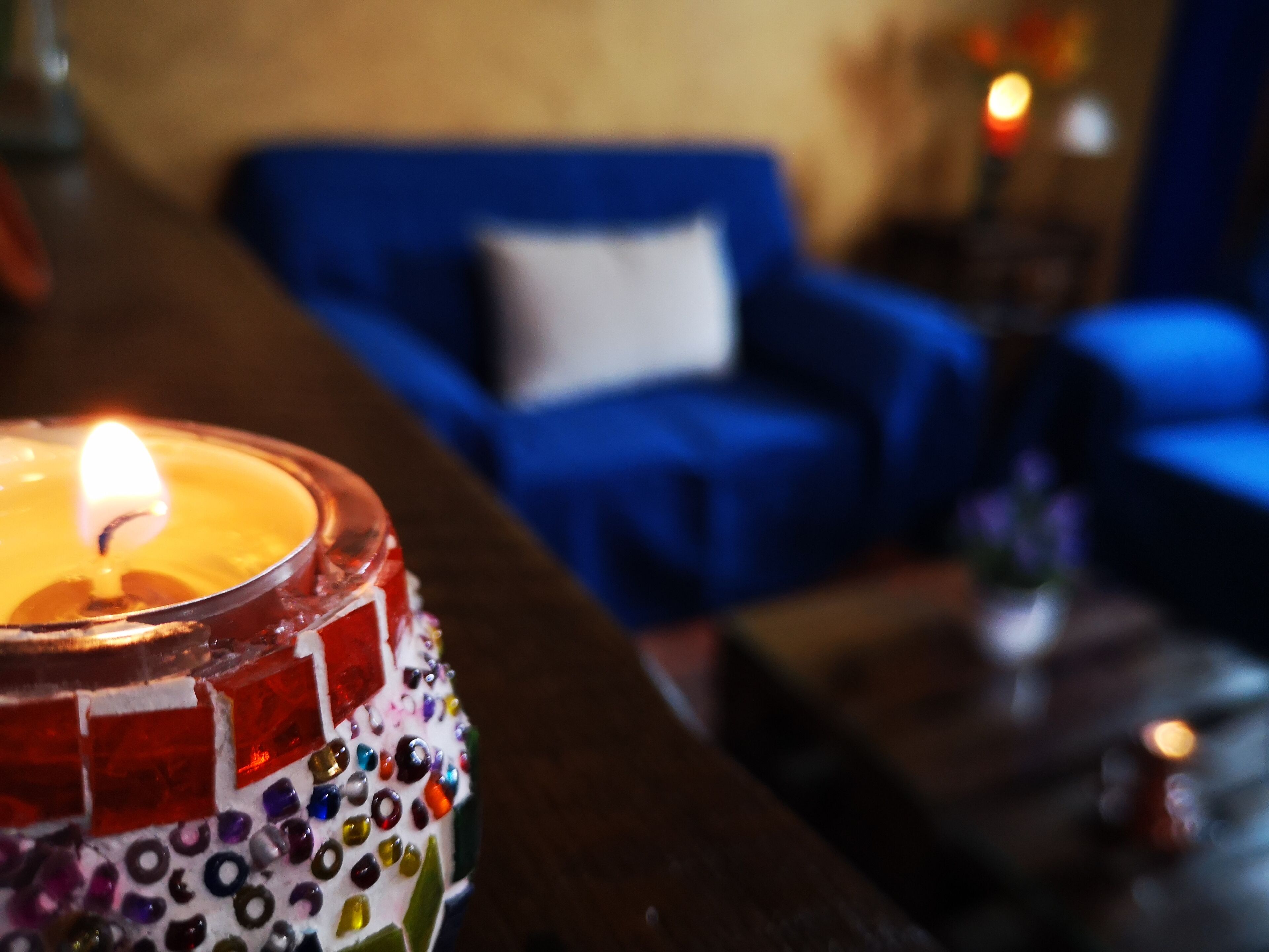 Un primo piano di una candela in un portacandela decorativo, con sullo sfondo sfocato un divano blu e un tavolino con un vaso.