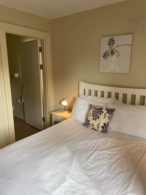 A neatly made bed with white linens and a single decorative pillow, a bedside lamp, and a door leading to a bathroom. - Dunkerron Woods 3 Bedroom 4024 (Kenmare)