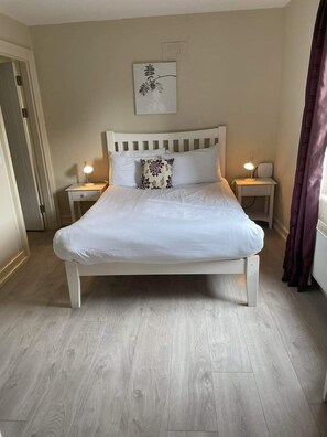 A neatly made bed with white bedding and a floral pillow, flanked by two bedside tables with lamps, in a room with light wooden flooring and a beige wall. - Dunkerron Woods 3 Bedroom 4027 (Kenmare)