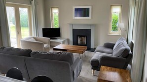 A modern living room with a sofa, armchair, TV, fireplace, and large windows. - Dunkerron Woods 3 Bedroom 4027 (Kenmare)