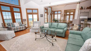 A spacious living room with a large window, a green sofa, a patterned armchair, and a glass coffee table. - OCEANFRONT, Ocean views, Rec Rm, Private Pool, Hot Tub, PI52, Coco Palms (Corolla)