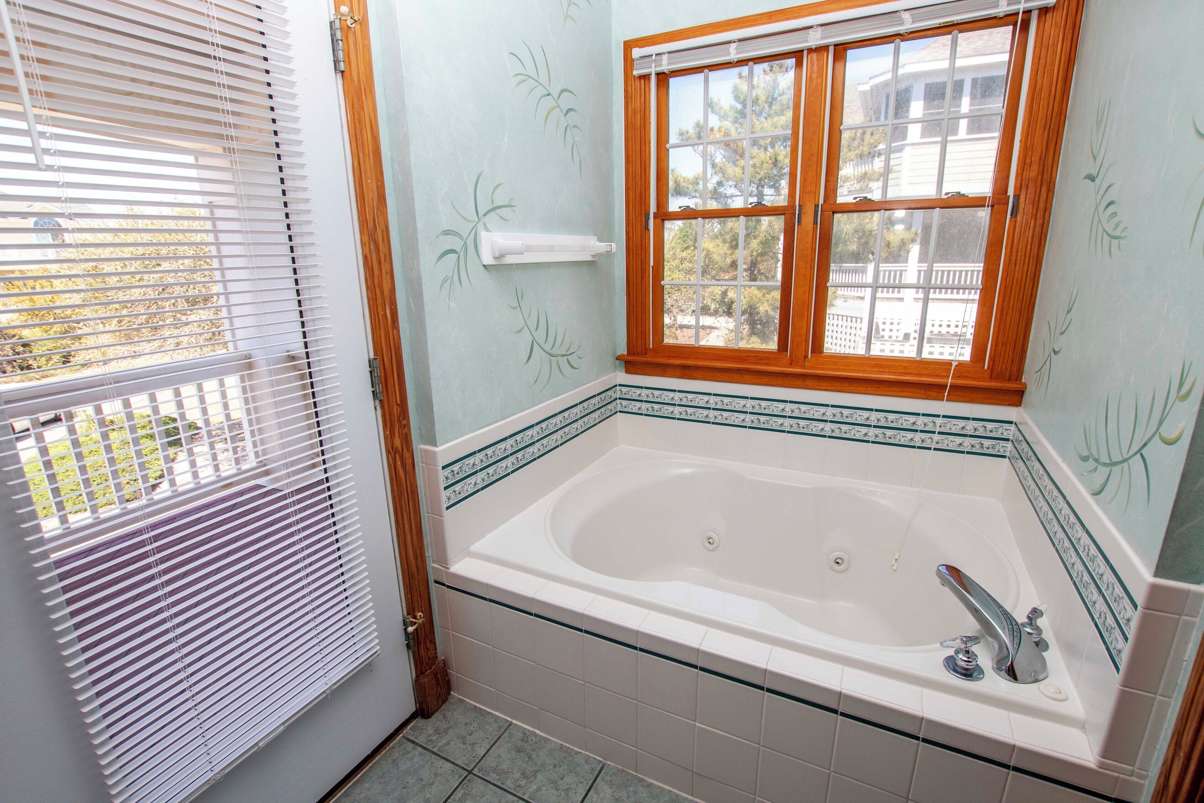A bathroom with a large bathtub, a window with wooden blinds, and a patterned tile border.
