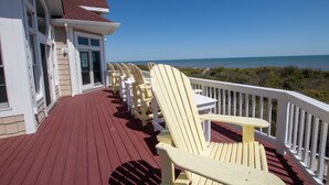 A wooden deck with red flooring and white railings, featuring several yellow Adirondack chairs and a small table. - OCEANFRONT, Ocean views, Rec Rm, Private Pool, Hot Tub, PI52, Coco Palms (Corolla)
