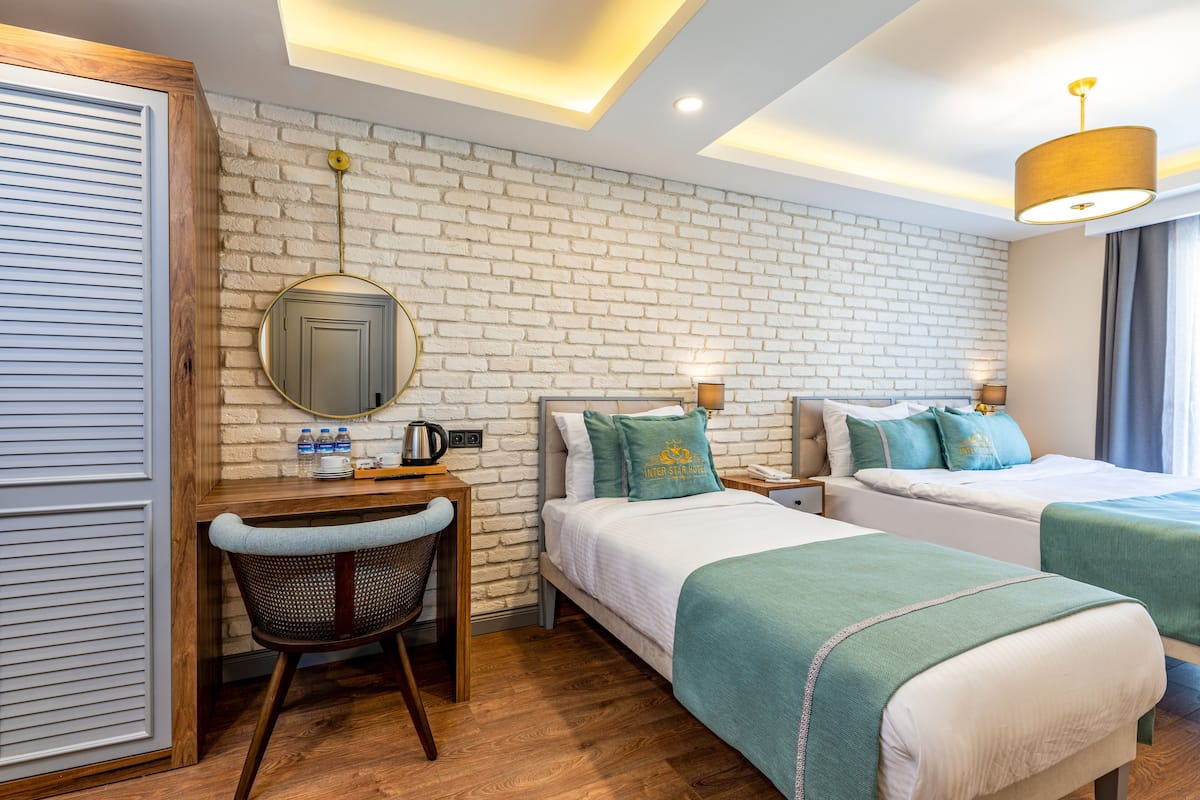 A hotel room with a brick wall, two beds, a desk with a chair, a mirror, and a hanging lamp.