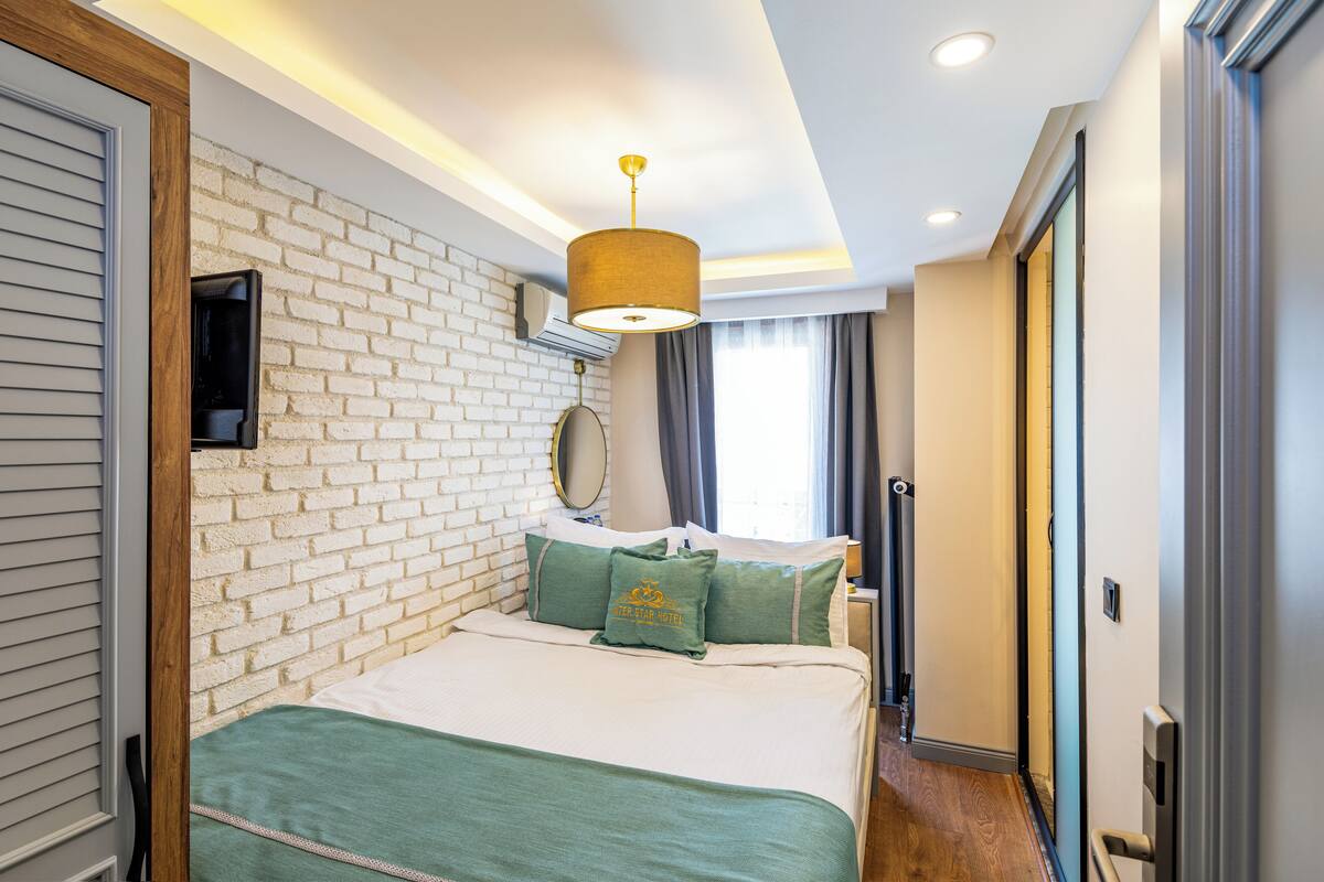 A hotel room with a brick wall, a bed with green bedding, a hanging lamp, and a window with curtains.