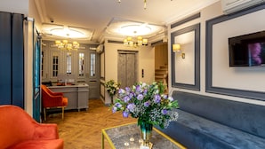 Un hall d’hôtel moderne avec une table basse en marbre, un fauteuil rouge, un canapé, un lustre et un téléviseur à écran plat fixé au mur.