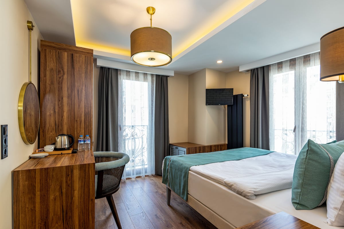 A hotel room with a wooden desk, a bed with green bedding, a television on the wall, and a balcony with curtains.
