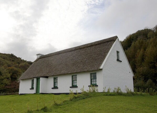A white building with a thatched roof, green doors, and windows, surrounded by greenery and hills. - Corofin 3 bed No 5 (Killinaboy)