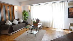 A living room with a sofa, a glass coffee table, a potted plant, and a large window with curtains. - Victoriei 7 - 2 bedroom apartment downtown (Sector 1)