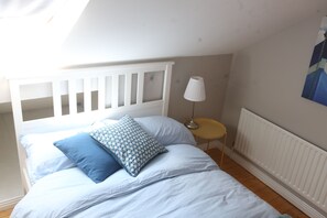 A bedroom with a white headboard, a bed with blue and white bedding, a small round table with a lamp, and a radiator on the wall.