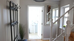 Un pasillo con un perchero, una planta en maceta, una puerta blanca que da al baño y una escalera con barandillas blancas.