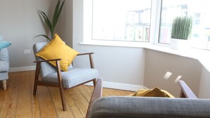 Una habitación con piso de madera, un sillón gris con un cojín amarillo, una planta en maceta y una ventana con vista a edificios.
