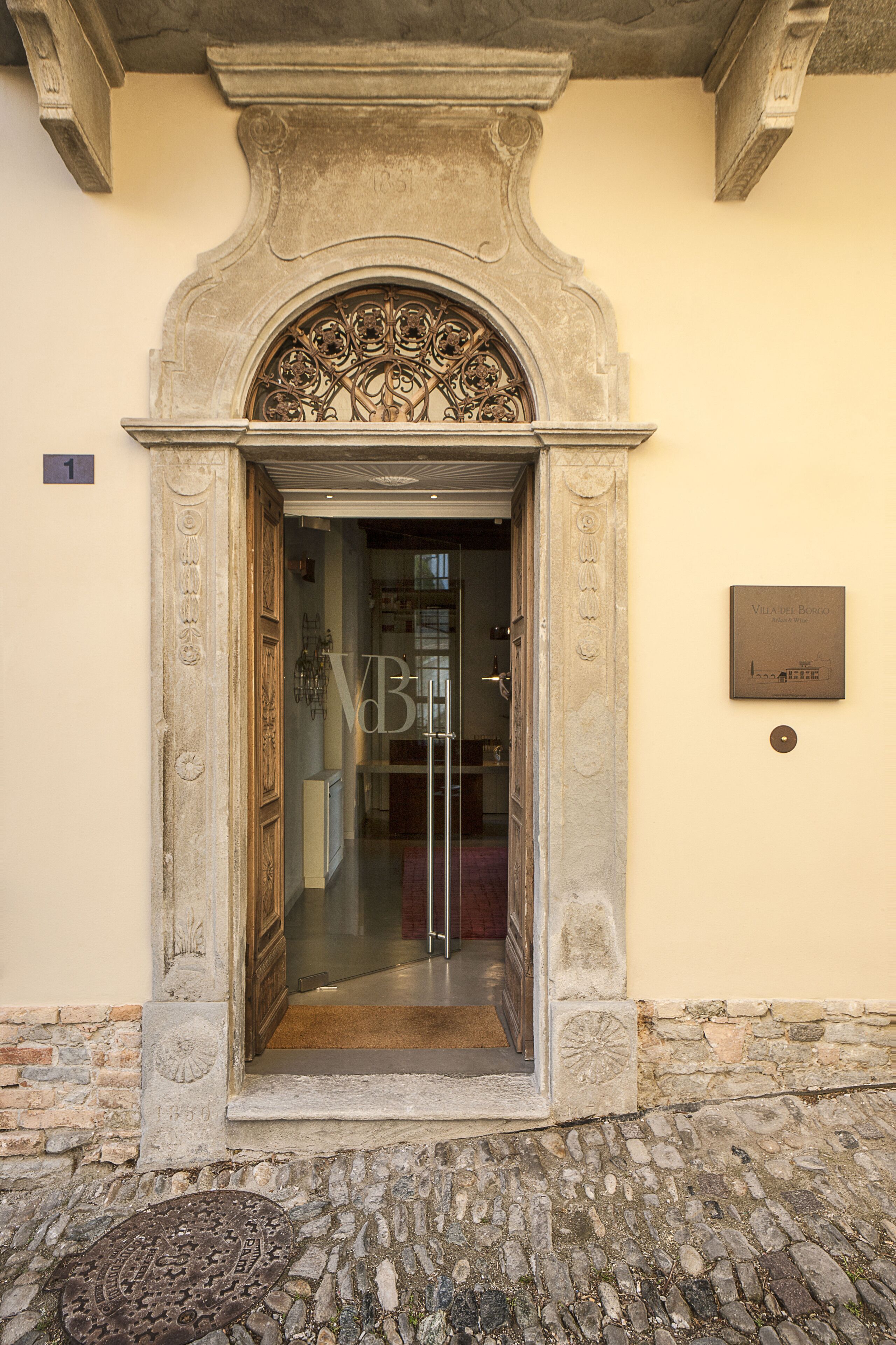 An ornate stone entrance with a glass door, a plaque on the wall, and a cobblestone ground.