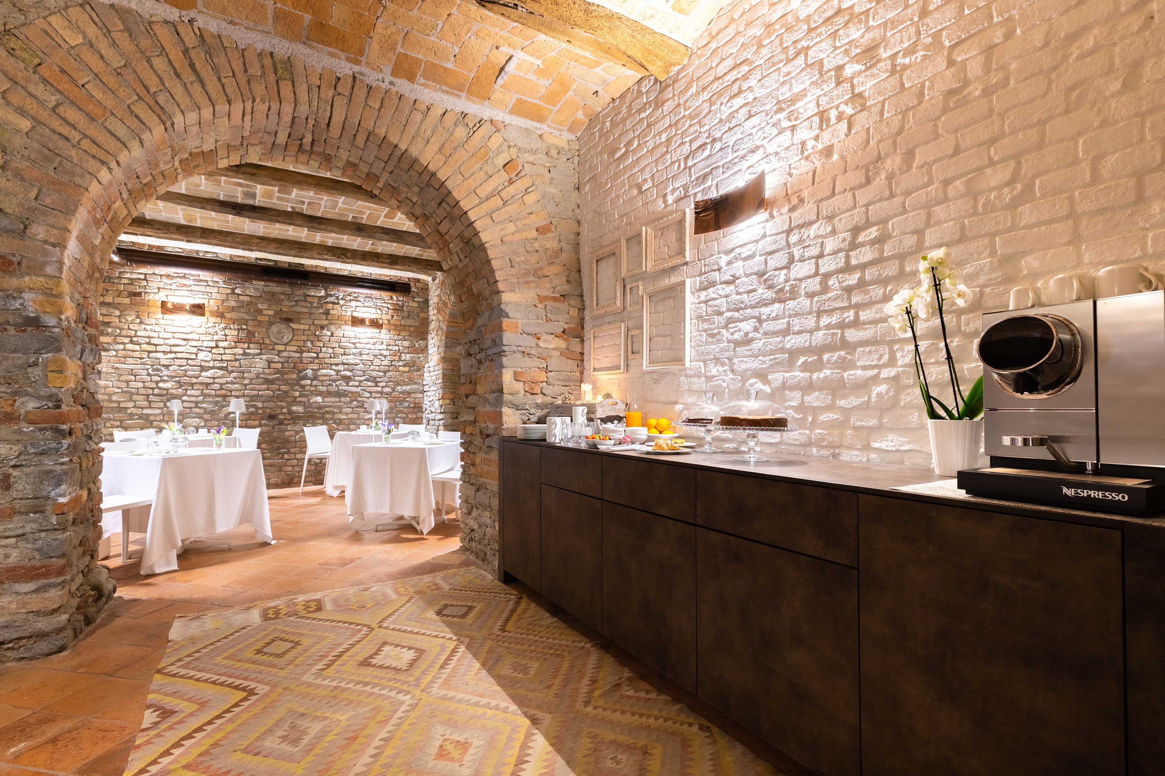 L’intérieur est de style rustique, avec des murs en pierre, une arche en briques et un comptoir où se trouve une machine à café. Les tables sont dressées pour le repas.