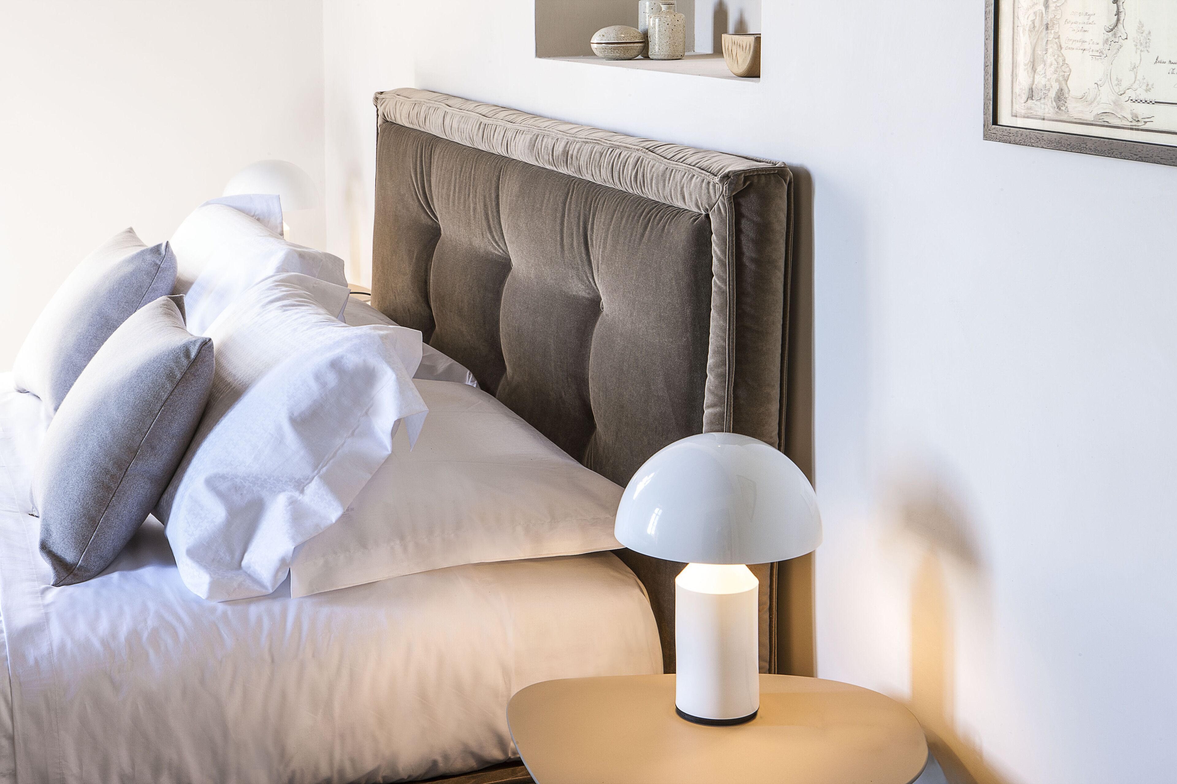 A neatly made bed with a tufted headboard, a bedside lamp, and a small shelf with decorative items.