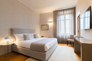 A modern hotel room with a large bed, bedside table, lamp, desk, chair, and a large window with curtains.