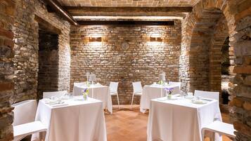A dining area with stone walls and brick ceiling, featuring round tables with white tablecloths and chairs.