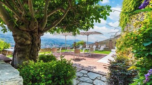 Un patio al aire libre con suelo de piedra, sillas blancas y sombrillas, con vistas a un paisaje pintoresco.