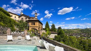 Zona de piscina con sillas de descanso, una piscina de piedra y vistas a una villa con torre del reloj.