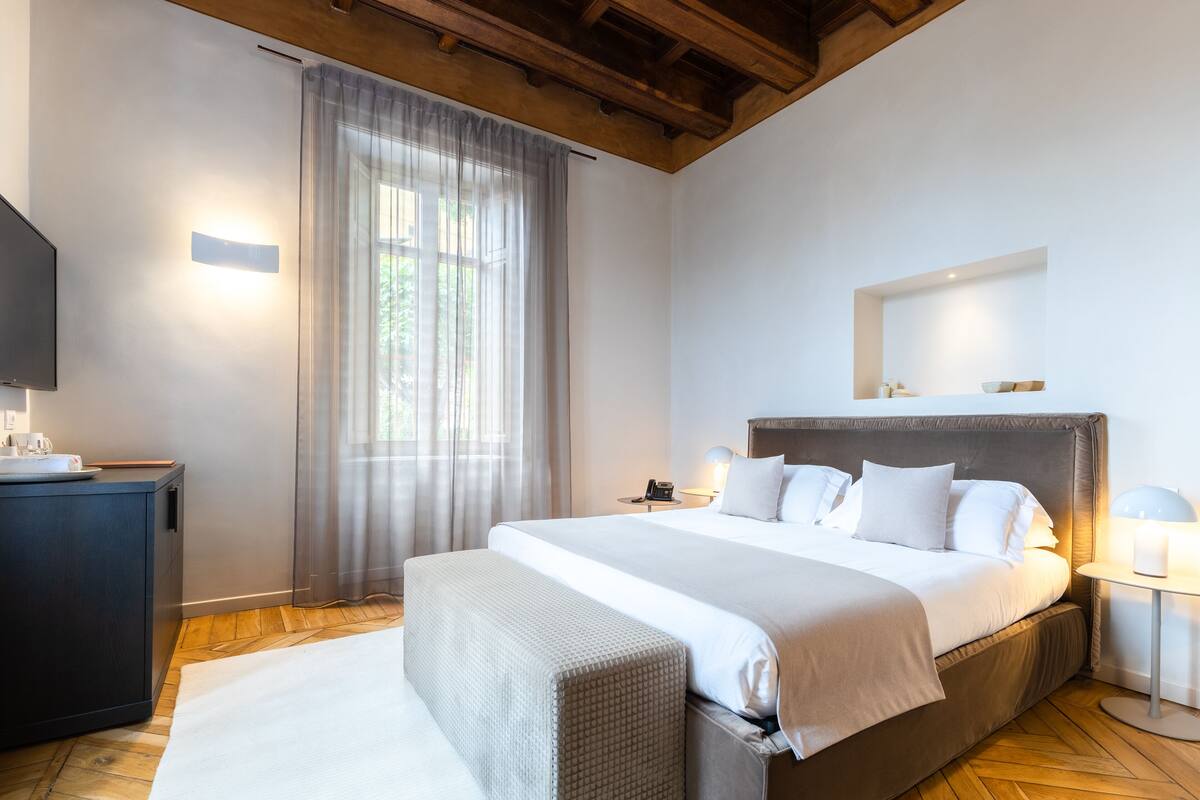 A modern hotel room with a large bed, a bedside table, a TV, and a window with sheer curtains.