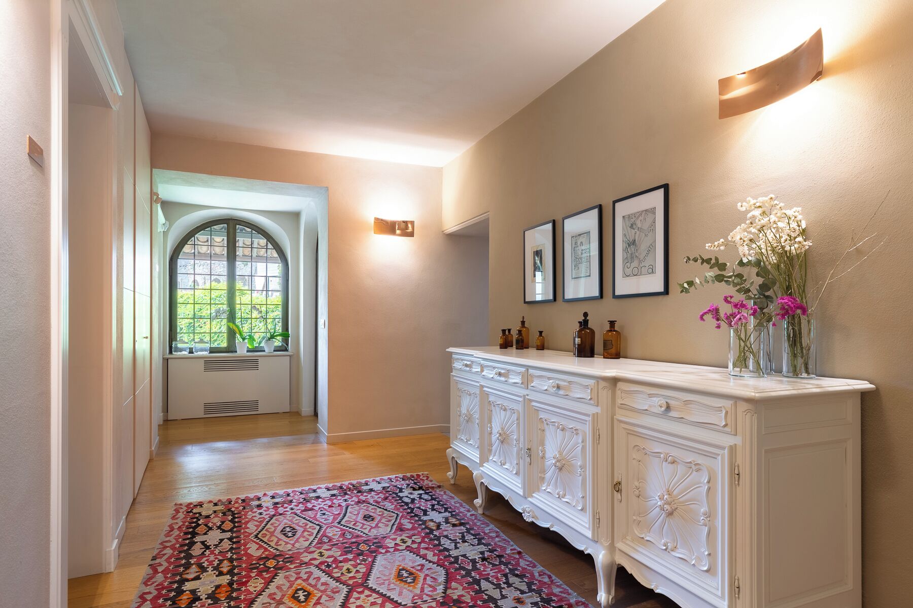 A hallway with a patterned rug, a white cabinet with decorative glass doors, framed pictures on the wall, and a window with a view of greenery.