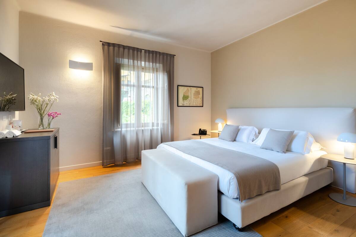 A modern hotel room with a large bed, a bedside table, a desk, and a window with curtains.