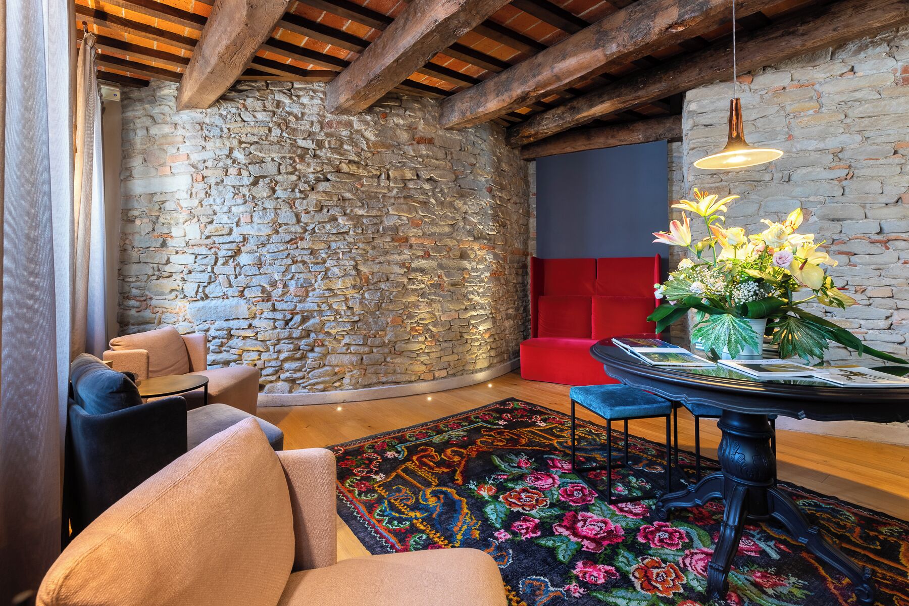 A room with a stone wall, wooden beams, a red sofa, a round table with a vase of flowers, and a colorful rug.