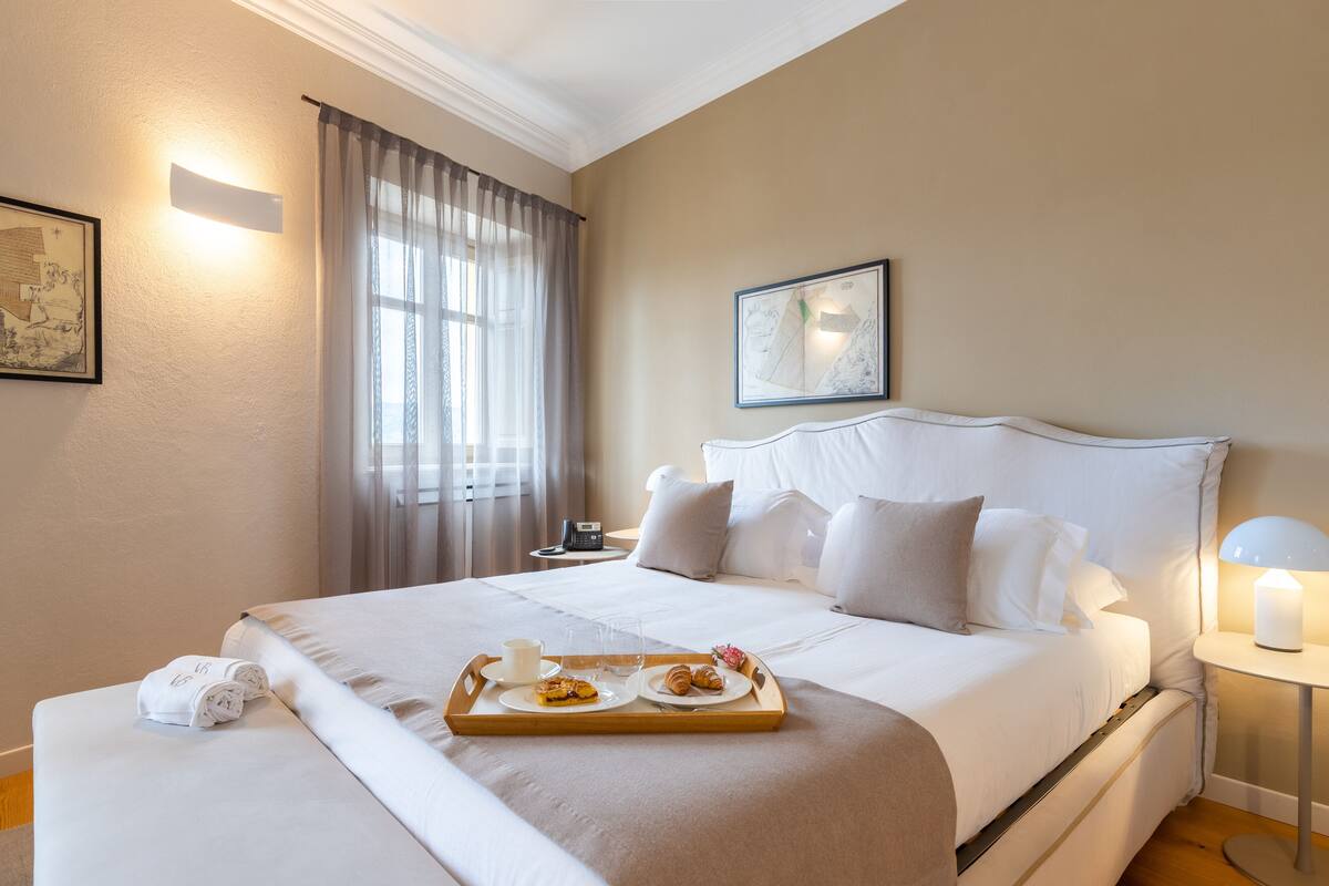 A neatly made bed with a tray of breakfast items, a bedside table with a lamp, and a large window with curtains.