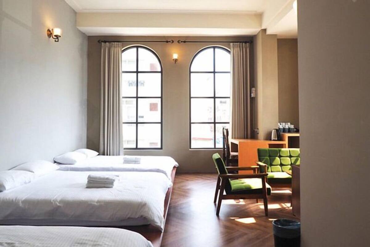 A hotel room with two beds, a desk, a chair, and large windows.