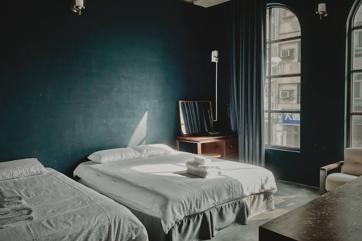 A hotel room with two beds, a desk, a chair, and a large window with a view of buildings outside.
