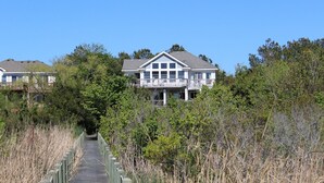 A large two-story house with a balcony, surrounded by dense vegetation and a wooden boardwalk leading to it.