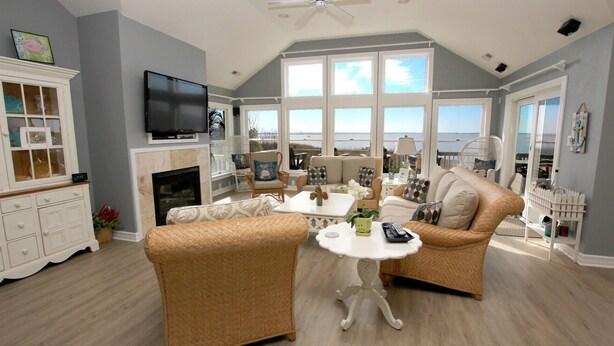 A spacious living room with a view of the ocean, wicker furniture, a fireplace, and a television.