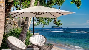 A beachside patio with wicker chairs and a white umbrella, overlooking the ocean. - Villa Gita Segara (Manggis)