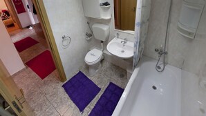 Een badkamer met toilet, wastafel en bad. Op de vloer liggen rode en paarse vloerkleden.