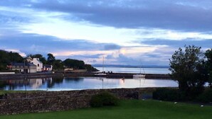 A coastal scene with a stone wall, grassy area, and buildings near the water. - Ballyvaughan No 7 (Ballyvaughan)