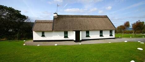 A traditional white cottage with a thatched roof, surrounded by a well-maintained lawn and a few scattered stones.