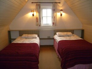 A small, cozy attic bedroom with two single beds, a window with curtains, and wall-mounted lamps.