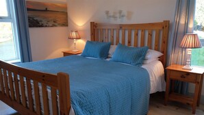 A bedroom with a wooden bed frame, a blue quilted bedspread, two bedside lamps, and a painting on the wall. - Ballyvaughan Cottage No 14 (Ballyvaughan)