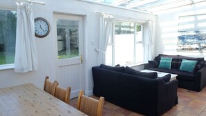 Una habitación luminosa con una mesa y sillas de madera, un sofá oscuro con cojines azul turquesa y una ventana con cortinas.